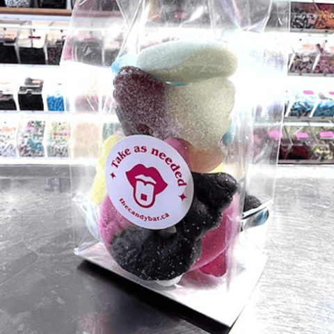 A clear plastic bag containing assorted BUBS candy pieces, with a round white sticker that reads 'Take as needed' and 'thecandybar.ca' attached in the center.
